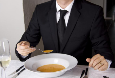 A man dressed in a black suit, white button up shirt, and a black tie sitting in a fancy restaurant with a bowl of bisque and a partially emptied glass of champagne.