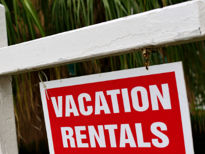 A white sign holder with a red "Vacation Rentals" sign hanging from it and palm trees in the background.