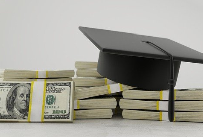 A pile of $100 bill bundles on a white table with a graduation cap sitting on top of the money.