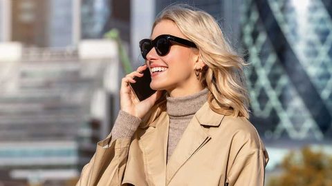 A blonde woman in a tan trench coat and sunglasses standing outside talking on her cell phone.