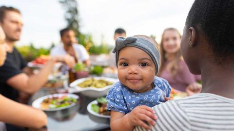A group of people sitting at a table (slightly out of focus) with a smiling baby looking at the camera.