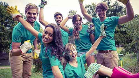 A group of young adults in matching green t-shirts all wearing gardening gloves celebrating outside in a wooded area.