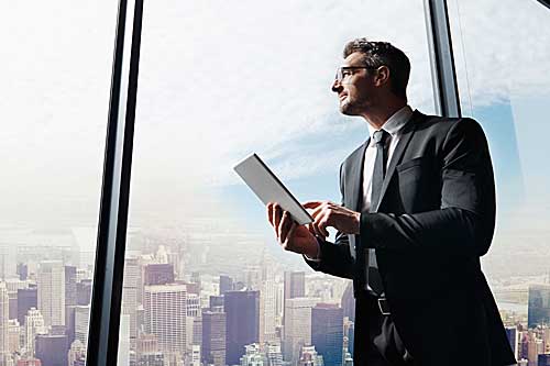A man holding a tablet and looking out the window over a city of high rise buildings.