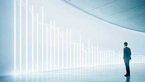 A lone man standing in front of a wall depicting a lighted line and bar graph of data.
