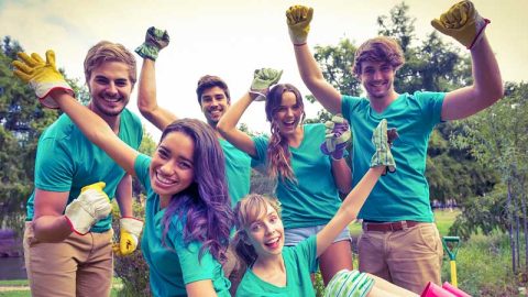A group of young adults in matching green t-shirts all wearing gardening gloves celebrating outside in a wooded area.